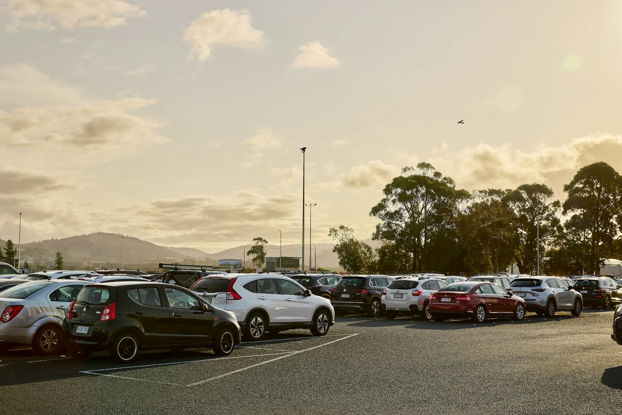 The sun casts shadows on the Hobart Airport long term carpark. A plane flies in the sky.