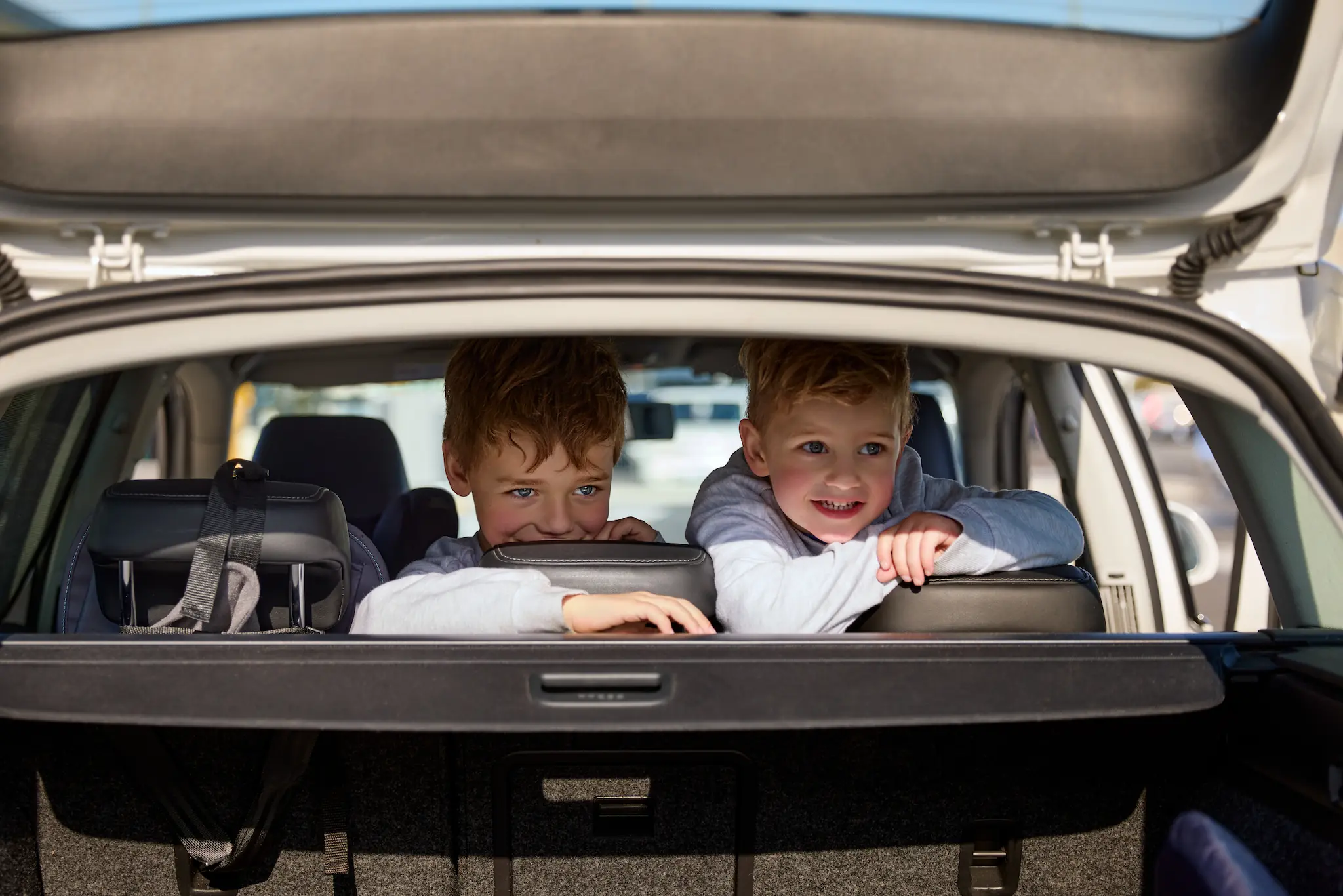 Two children smile at the camera over the backs of their seats through the car's open boot
