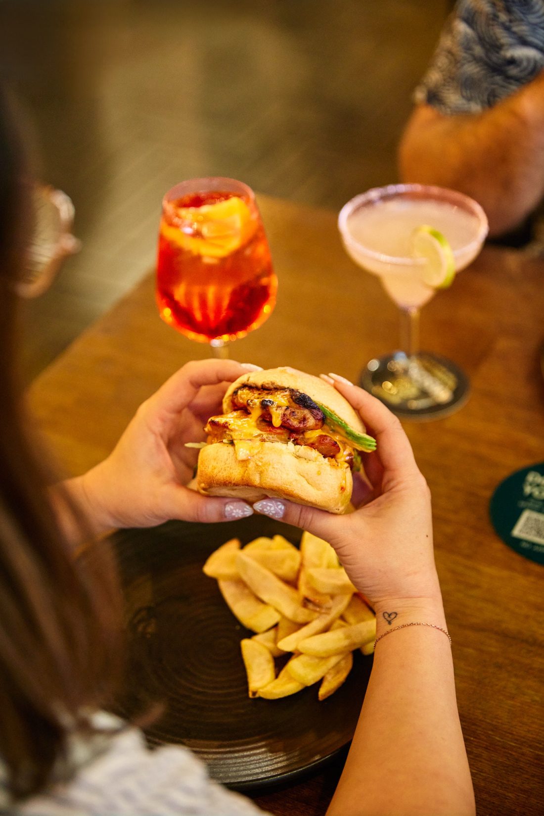 Two hands holding on to a juicy burger with a plate of chips under, two cocktail glasses on the table.
