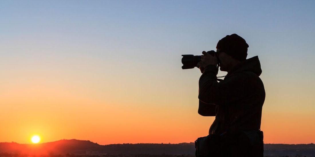 Silhouette of a person in a hat and jacket taking a photo of the sunset, with a clear sky showing an orange and blue gradient over distant hills.